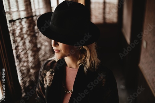 Charming pensive young female in elegant vintage clothes and hat standing in front of window in dark room