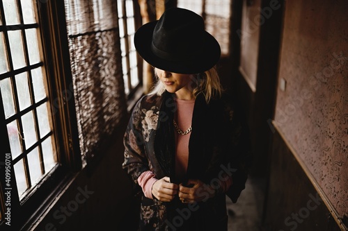 Stylish female in elegant vintage outfit and hat standing in narrow hallway of old house and looking at mirror