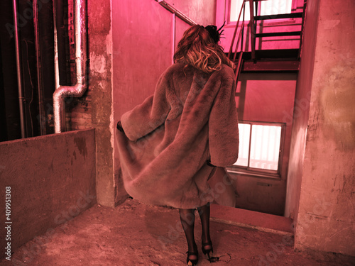Back view stylish drag queen in elegant dress and fur coat looking away while standing near staircase under red light inside grungy building