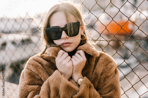 Young determined female in sunglasses and warm jacket standing near net fence and looking at camera photo