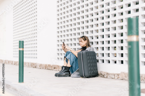 Side view of female tourist with luggage sitting alone on pavement and browsing mobile phone while getting lost in city
