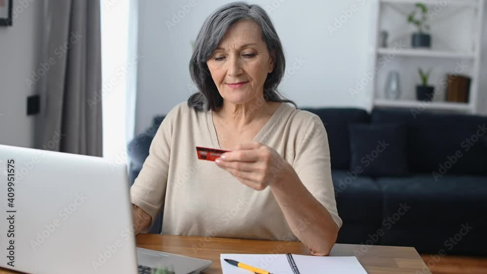 Beautiful senior woman holding a credit card and using a laptop for making a purchase on the Internet, transferring money, e-banking sitting at the table at home Beautiful senior woman holding a credit card and using a laptop for making a purchase on the Internet, transferring money, e-banking sitting at the table at home