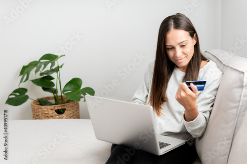Female buyer sitting on sofa at home and making payment with plastic card while shopping online via laptop