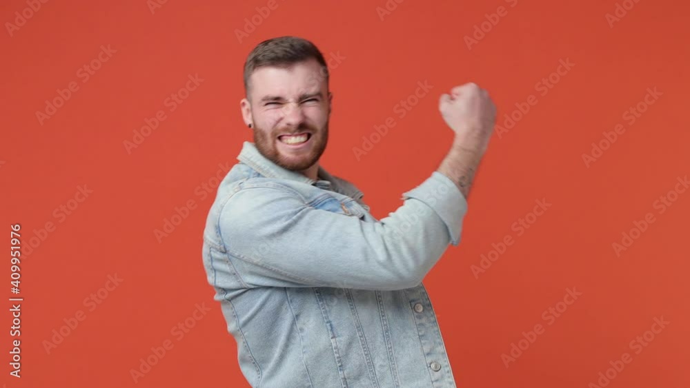 Happy overjoyed bearded young man 20s in casual white t-shirt denim jacket isolated on orange background in studio. People lifestyle concept. Doing winner gesture celebrating clenching fists say yes Happy overjoyed bearded young man 20s in casual white t-shirt denim jacket isolated on orange background in studio. People lifestyle concept. Doing winner gesture celebrating clenching fists say yes