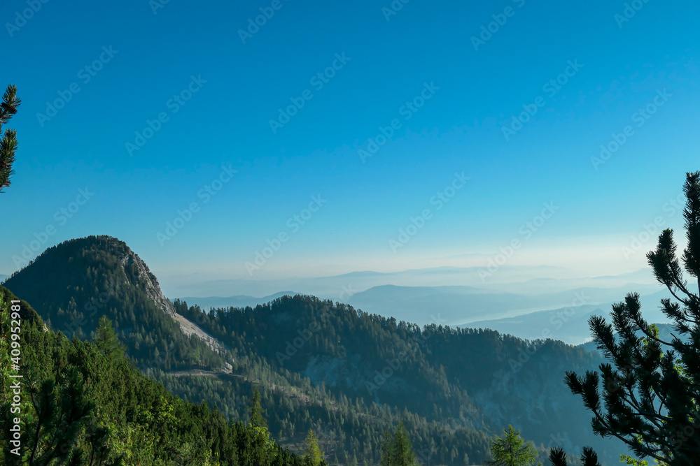 A panoramic view on the Alps from the top of Mittagskogel in Austria. Clear and sunny day. Sharp peaks around. A bit of haze in the valley. Outdoor activity. Alpine mountain chains in the back. Calm