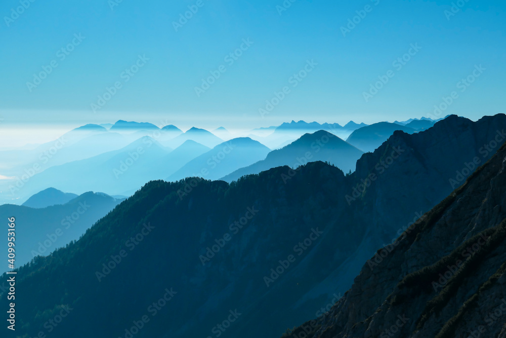 A close up view on the Alpine chains shrouded in the morning fog, seen from the top of Mittagskogel in Austria. Clear and sunny day. Sharp peaks around. Sun is shining above the high peaks. Serenity