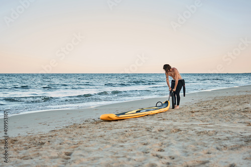 Man in wetsuit inflating paddle board at the seashore on sandy beach