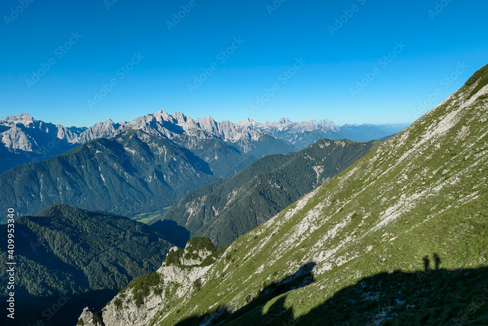 A panoramic view on the Alps from the top of Mittagskogel in Austria There is a shadow of two people standing at the top. Clear and sunny day. Sharp peaks around. A bit of haze in the valley. Fun A panoramic view on the Alps from the top of Mittagskogel in Austria There is a shadow of two people standing at the top. Clear and sunny day. Sharp peaks around. A bit of haze in the valley. Fun