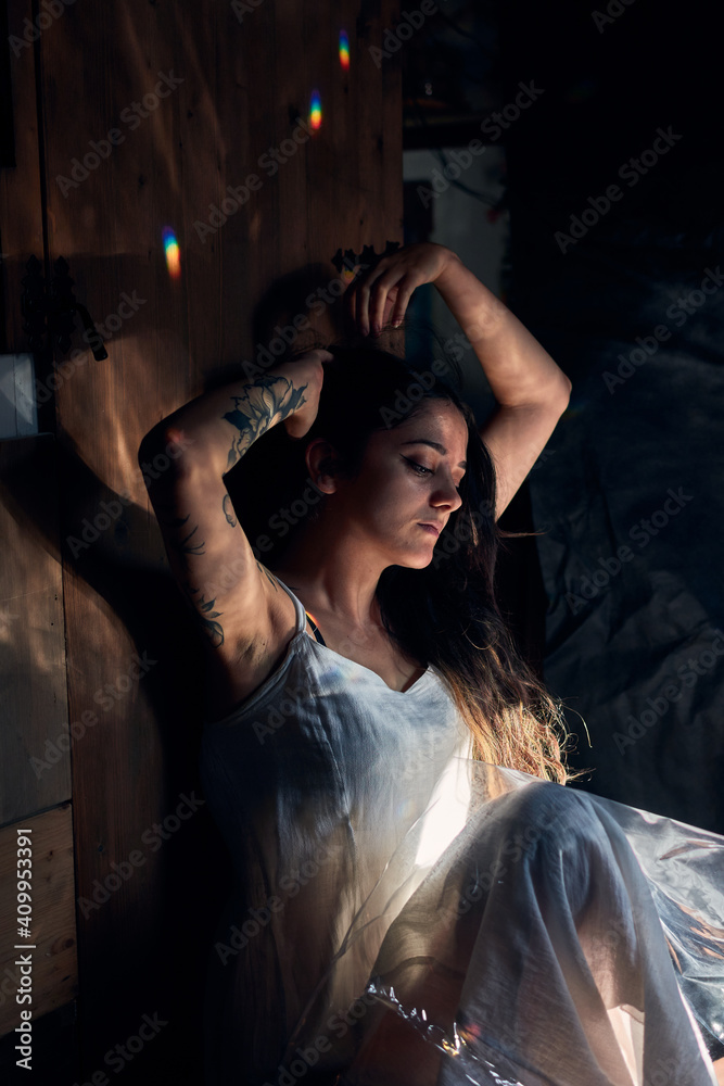 Gentle female in white maxi dress sitting on floor near wooden wall and touching hair in dark room Gentle female in white maxi dress sitting on floor near wooden wall and touching hair in dark room