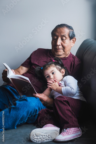 Senior ethnic grandfather reading interesting story in book for cute little girl while relaxing together on sofa at home