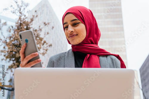 Cheerful Arab female freelancer in red hijab sitting with laptop on bench in city and browsing smartphone while working on startup project remotely photo