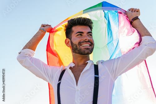 Low angle of expressive homosexual male standing on street with bright rainbow flag and cheerfully screaming