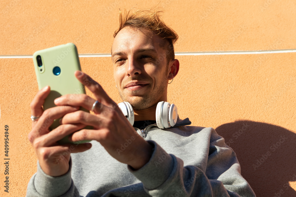 © David Munoz/ADDICTIVE STOCK - Low angle of positive young guy in casual outfit with headphones on neck sitting near stone wall and using mobile phone in sunny day in city © David Munoz/ADDICTIVE STOCK - Low angle of positive young guy in casual outfit with headphones on neck sitting near stone wall and using mobile phone in sunny day in city