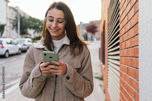 Positive female millennial chatting on social media with friends via smartphone while standing on street in city photo