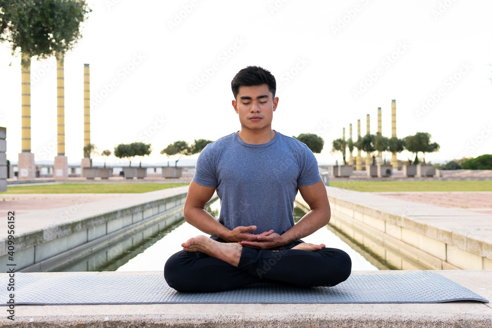 Flexible Asian male sitting on mat in Padmasana and meditating with closed eyes while practicing yoga in park