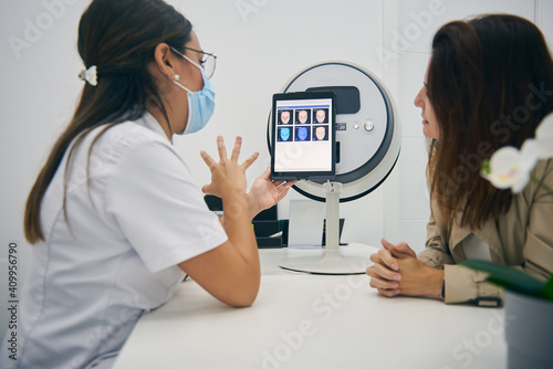 Side view of cosmetologist and customer sitting at table with facial skin diagnostics equipment while looking at tablet and discussing results of analysis in beauty clinic photo