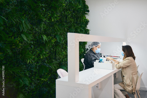 Side view of cheerful manicure master using nail file while sitting at table with female client in modern beauty salon during coronavirus pandemic