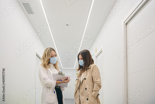 Female client in mask talking to beautician standing with clipboard in white corridor of modern beauty center photo