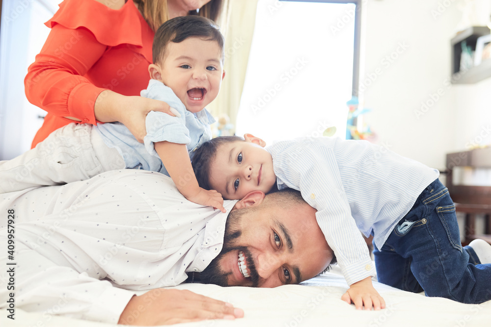 Positive Hispanic couple with cute little sons gathering on bed and looking at camera while enjoying pastime at home Positive Hispanic couple with cute little sons gathering on bed and looking at camera while enjoying pastime at home