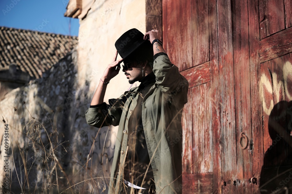 Side view of unrecognizable male traveler in trendy coat putting on black hat while standing near weathered shabby rural building in countryside