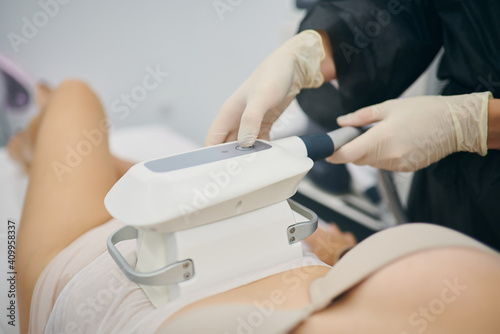 Cropped unrecognizable slim female client lying on table and getting anti cellulite massage of belly with LPG machine while looking at professional masseuses photo