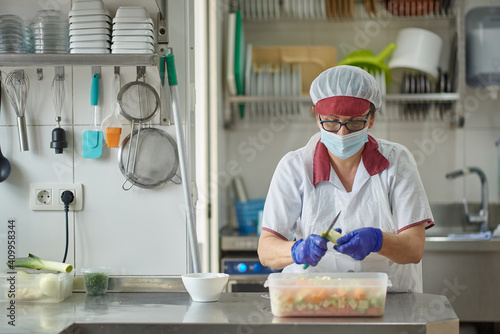 Female cook in white uniform and protective mask and gloves cutting fresh vegetables while preparing food in hospital kitchen during coronavirus pandemic photo