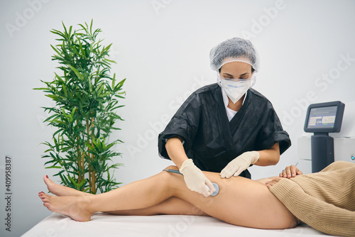 Professional masseuse doing anti cellulite massage on legs of female client while using special machine in modern beauty clinic photo