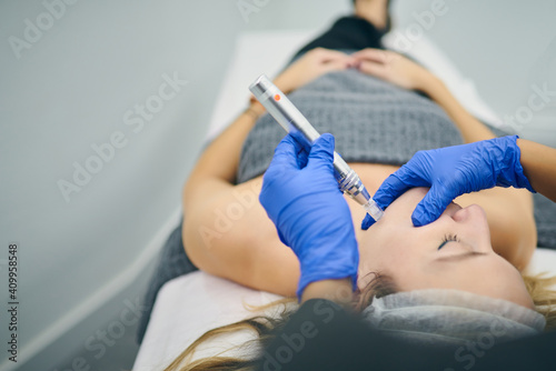 Cropped unrecognizable cosmetologist in uniform using derma pen with micro needles and cleaning face of client in beauty clinic photo