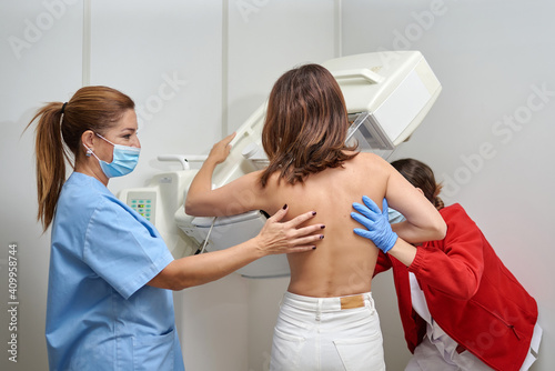 Female medics in masks and uniform preparing woman with naked torso for chest radiology while standing in medical room in clinic photo