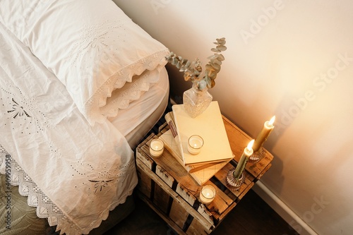 Fragment of interior of bedroom with rustic wooden handmade bedside table with dry plants in glass vase placed on stack of old books near burning candles