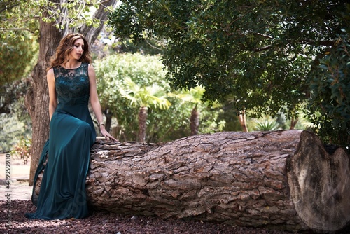 Full body of dreamy female wearing maxi emerald dress sitting on tree trunk in woods and looking down photo