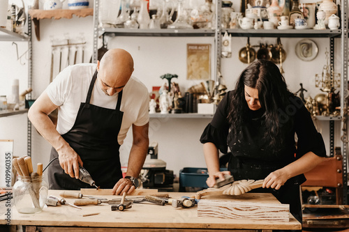 Concentrated mature artisan man and woman with professional tools creating wooden details while working together in art workshop photo