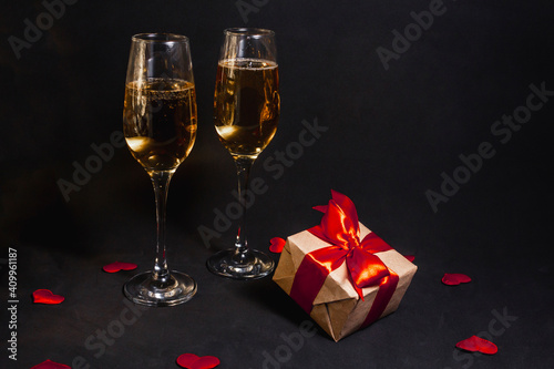 two glasses of champagne stand in the dark, next to a gift box tied with a red ribbon, and confetti made of hearts © Anastasiya Famina
