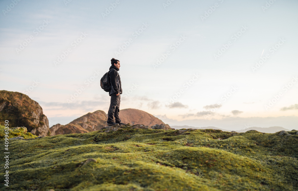 Side view of lonely male explorer with backpack standing on top of green hill and contemplating nature while traveling through mountainous terrain Side view of lonely male explorer with backpack standing on top of green hill and contemplating nature while traveling through mountainous terrain
