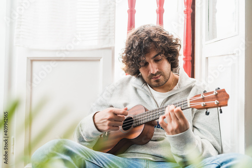 Thoughtful young curly haired ethnic musician in casual wear performing music on ukulele guitar against white wall photo