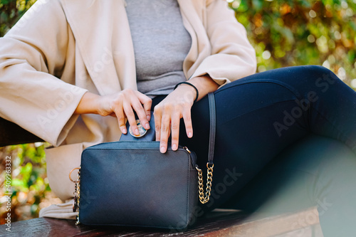 Side view of unrecognizable crop female taking mobile phone from handbag while sitting on wooden bench in park photo