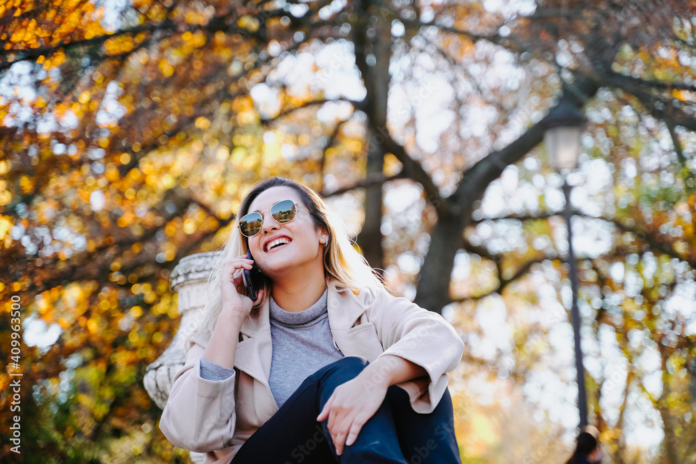 Low angle of content female sitting in autumn park and talking on smartphone while enjoying conversation and looking away Low angle of content female sitting in autumn park and talking on smartphone while enjoying conversation and looking away