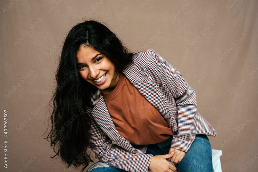 Stylish cheerful ethnic female model in trendy smart casual outfit sitting on chair and looking confidently at camera Stylish cheerful ethnic female model in trendy smart casual outfit sitting on chair and looking confidently at camera