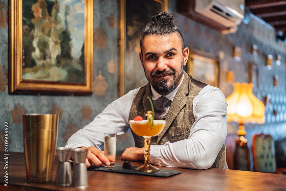 Adult elegant man smiling looking at camera driking liquor near tropical cocktail while sitting at counter and resting in bar Adult elegant man smiling looking at camera driking liquor near tropical cocktail while sitting at counter and resting in bar