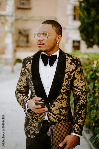 Side view of rich African American male in posh tuxedo standing in street while browsing on smartphone looking away