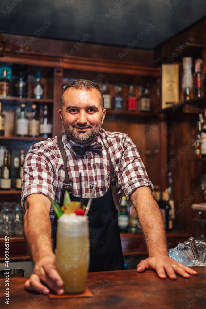 Happy male mixologist putting glass of cold exotic cocktail with fruits and ice on counter during work in bar looking at camera Happy male mixologist putting glass of cold exotic cocktail with fruits and ice on counter during work in bar looking at camera