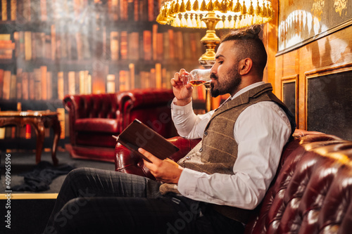 Side view of relaxed bearded male using tablet and enjoying liquor while resting on sofa in elegant pub photo