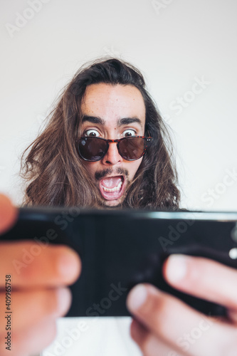 Overwhelmed young long haired hipster guy in sunglasses looking at screen of smartphone with astonished face expression while getting incredible news