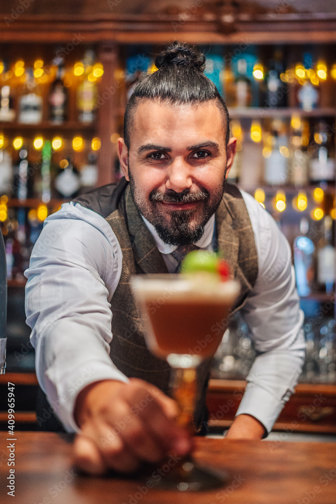 Happy bearded male mixologist putting glass of cold exotic cocktail with fruits and ice on counter during work in bar looking at camera Happy bearded male mixologist putting glass of cold exotic cocktail with fruits and ice on counter during work in bar looking at camera