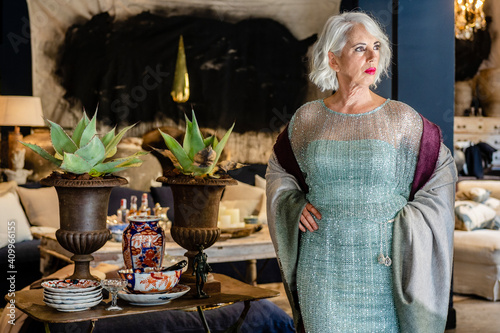 Confident mature female wearing sparkling glamour dress standing with hands on waist in luxury room in house and looking away