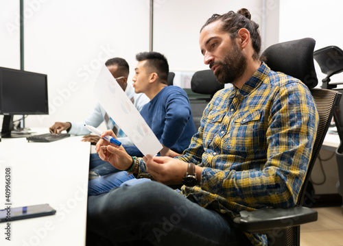 Side view of focused bearded male analyst manager in casual outfit examining paper with diagrams while working in modern workspace with colleagues photo