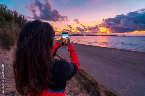 Back view of unrecognizable female traveler with long dark hair in casual clothes taking picture of amazing sunset over sea with sandy beach on smartphone during holidays in Cantabria