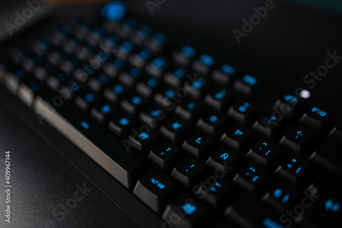 From above of black keyboard with blue illuminated letters placed on desk near computer monitor