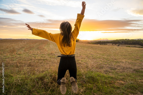 Back view os unrecognizable carefree female in casual outfit in moment of jumping above ground in meadow on background of sunset in rural area photo