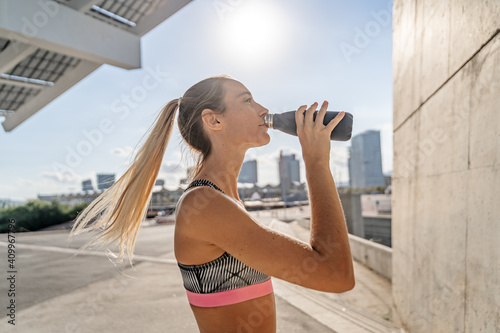 Side view of thirsty female athlete drinking fresh water from bottle while standing on street and having break during training